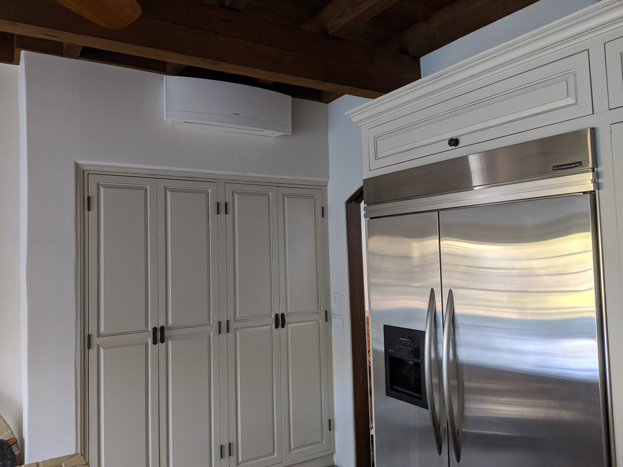 White wall-mounted mini-split air conditioner installed under a dark wooden ceiling above a white wardrobe in the corner of the room.