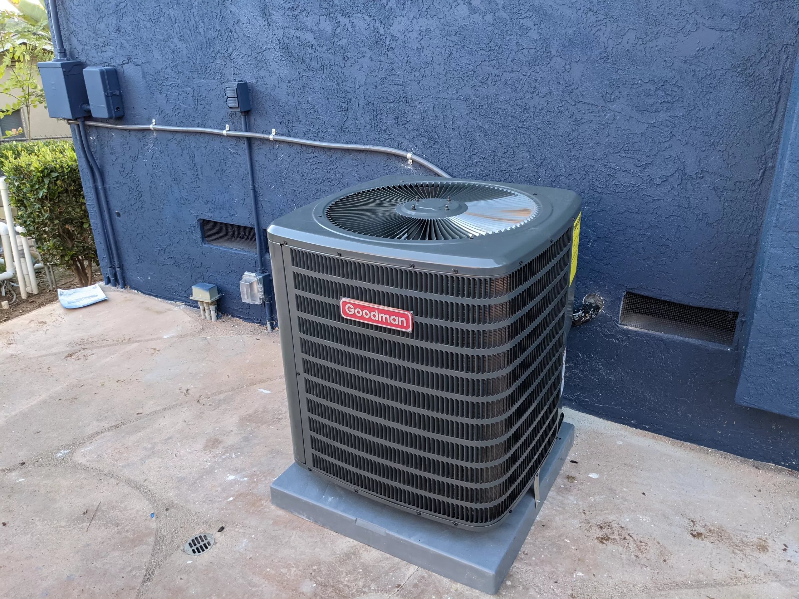 Goodman heat pump installed near the blue wall on the outside of the house.